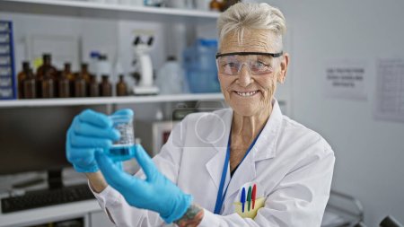 Téléchargez les photos : Femme scientifique aux cheveux gris âgée et confiante, souriante alors qu'elle travaille méticuleusement à une expérience délicate dans le laboratoire animé, éprouvette à la main - en image libre de droit