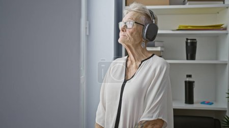 Foto de Relajado senior gris de pelo mujer de negocios trabajador se sumerge en la música, mirando tirar ventana de la oficina - Imagen libre de derechos