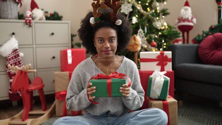Photo for African american woman wearing reindeer ears holding christmas gift at home - Royalty Free Image