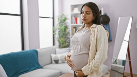 Foto de Mujer embarazada joven emocional, tocando suavemente su vientre, profundamente en el pensamiento en casa, en un fondo de la sala de estar. - Imagen libre de derechos