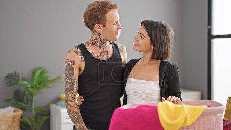 Téléchargez les photos : Beau couple étreignant et se regardant dans le panier avec des vêtements souriant à la buanderie - en image libre de droit