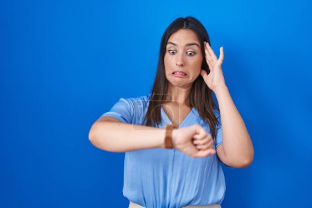 Téléchargez les photos : Jeune femme brune debout sur fond bleu regardant le temps de la montre inquiet, peur de se faire tard - en image libre de droit