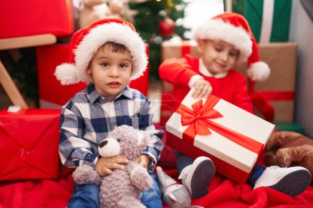 Téléchargez les photos : Deux enfants tenant cadeau et ours en peluche assis sur le sol près de l'arbre de Noël à la maison - en image libre de droit