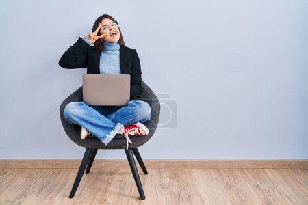 Photo for Young hispanic woman sitting on chair using computer laptop doing peace symbol with fingers over face, smiling cheerful showing victory - Royalty Free Image