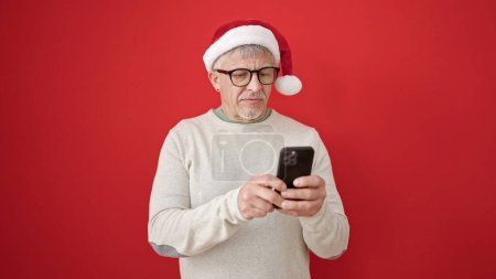 Téléchargez les photos : Homme moyen âge aux cheveux gris portant un chapeau de Noël en utilisant un smartphone sur fond rouge isolé - en image libre de droit