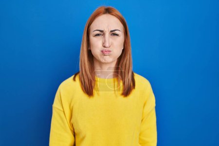 Foto de Young woman standing over blue background puffing cheeks with funny face. mouth inflated with air, crazy expression. - Imagen libre de derechos