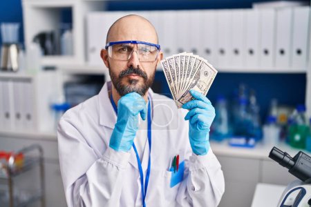 Photo for Young hispanic man working at scientist laboratory holding money serious face thinking about question with hand on chin, thoughtful about confusing idea - Royalty Free Image