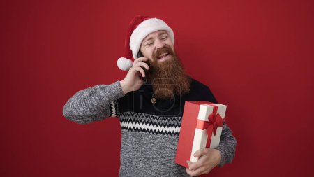 Téléchargez les photos : Jeune rousse homme tenant cadeau de Noël parler sur smartphone sur fond rouge isolé - en image libre de droit
