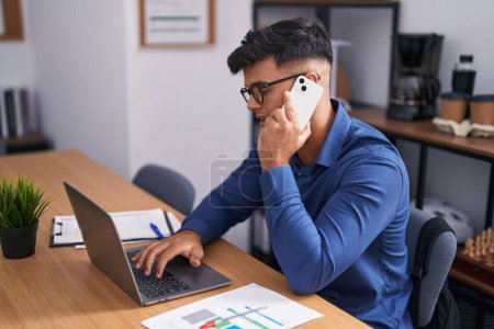 Foto de Young hispanic man business worker using laptop talking on smartphone at office - Imagen libre de derechos