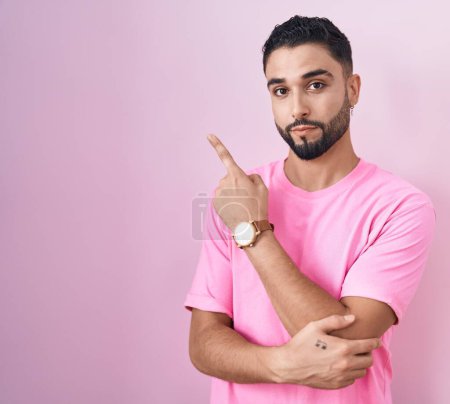 Photo for Hispanic young man standing over pink background pointing with hand finger to the side showing advertisement, serious and calm face - Royalty Free Image