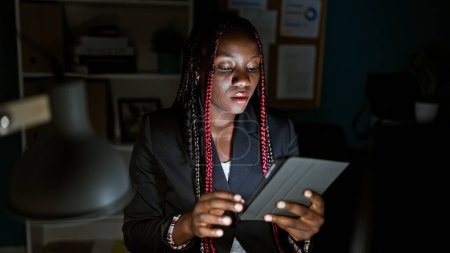 Téléchargez les photos : Travailleuse afro-américaine concentrée au bureau, surveille la lueur, les tresses et la veste sombre, les doigts agiles sur le pavé tactile et l'ordinateur, ce qui illustre le succès élégant de l'entreprise - en image libre de droit