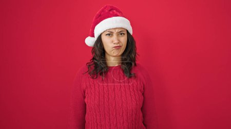 Photo for Young beautiful hispanic woman standing looking upset wearing christmas hat over isolated red background - Royalty Free Image