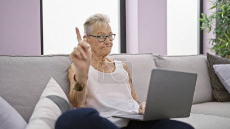 Téléchargez les photos : Femme âgée aux cheveux gris sérieux disant non avec un geste ferme du doigt tout en utilisant l'ordinateur portable sur un canapé confortable à la maison - en image libre de droit