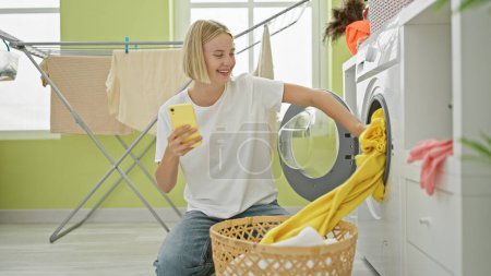 Téléchargez les photos : Jeune femme blonde utilisant smartphone laver les vêtements en souriant à la buanderie - en image libre de droit