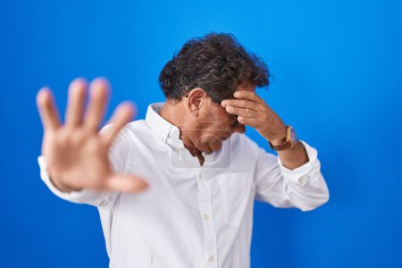 Photo for Middle age hispanic man standing over blue background covering eyes with hands and doing stop gesture with sad and fear expression. embarrassed and negative concept. - Royalty Free Image