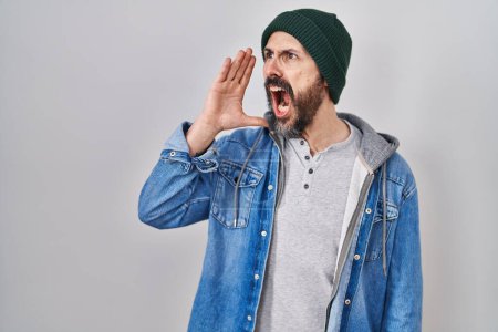 Foto de Joven hombre hispano con tatuajes con gorra de lana gritando y gritando alto a lado con la mano en la boca. concepto de comunicación. - Imagen libre de derechos