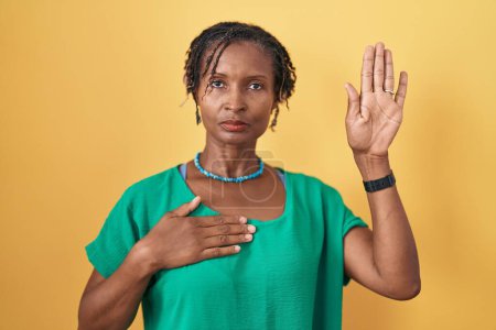 Téléchargez les photos : Femme africaine avec dreadlocks debout sur fond jaune jurant avec la main sur la poitrine et la paume ouverte, faire un serment de fidélité promesse - en image libre de droit