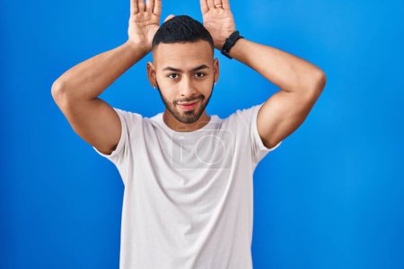Foto de Young hispanic man standing over blue background doing bunny ears gesture with hands palms looking cynical and skeptical. easter rabbit concept. - Imagen libre de derechos
