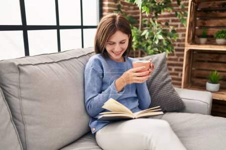 Foto de Joven mujer rubia bebiendo café libro de lectura en casa - Imagen libre de derechos