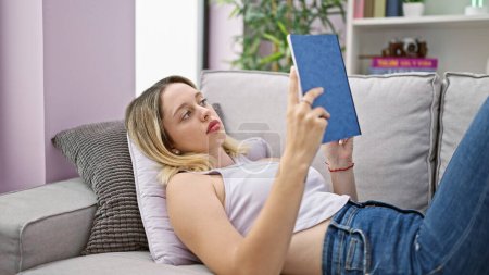 Téléchargez les photos : Young blonde woman reading book lying on sofa at home - en image libre de droit