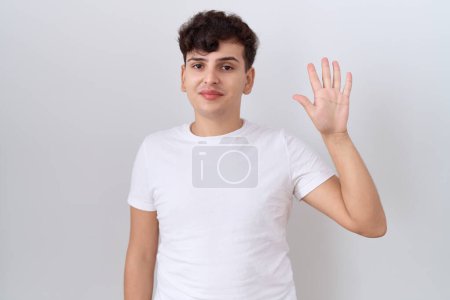 Foto de Joven hombre no binario con camiseta blanca casual mostrando y apuntando hacia arriba con los dedos número cinco mientras sonríe confiado y feliz. - Imagen libre de derechos