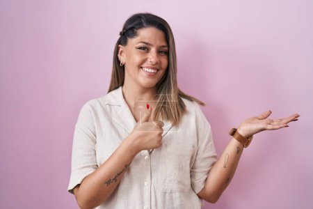 Foto de Mujer rubia de pie sobre fondo rosa mostrando la palma de la mano y haciendo el gesto bien con los pulgares hacia arriba, sonriendo feliz y alegre - Imagen libre de derechos
