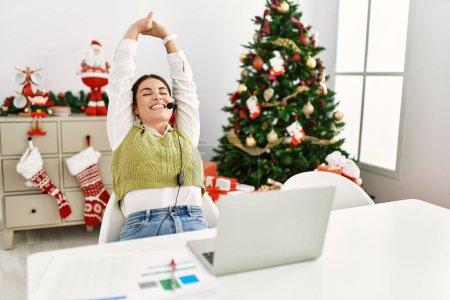 Photo for Young beautiful hispanic woman call center agent relaxed sitting by christmas tree at home - Royalty Free Image