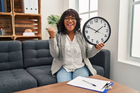 Photo for Hispanic woman working at therapy office holding clock screaming proud, celebrating victory and success very excited with raised arms - Royalty Free Image