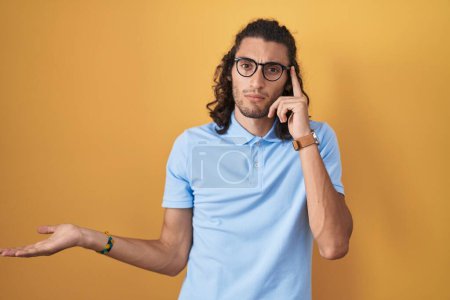 Young hispanic man standing over yellow background confused and annoyed with open palm showing copy space and pointing finger to forehead. think about it. 