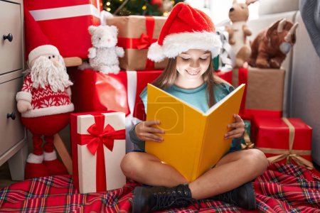 Photo for Adorable hispanic girl reading book sitting on floor by christmas tree at home - Royalty Free Image