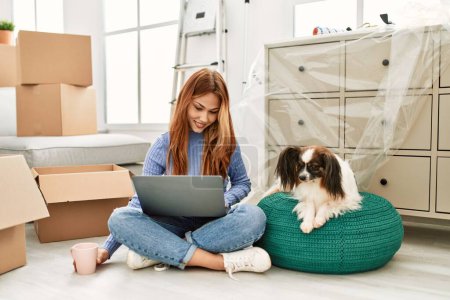Téléchargez les photos : Jeune femme caucasienne utilisant ordinateur portable assis sur le sol avec chien à la nouvelle maison - en image libre de droit