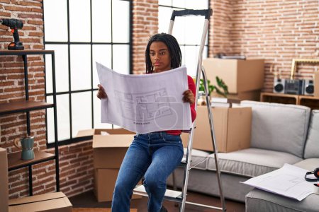 Photo for Young african american with braids standing on new home looking at blueprints relaxed with serious expression on face. simple and natural looking at the camera. - Royalty Free Image
