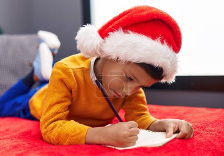 Photo for Adorable hispanic boy wearing christmas hat writing santa claus letter at home - Royalty Free Image