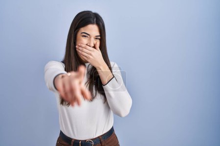 Téléchargez les photos : Jeune femme brune debout sur fond bleu se moquant de vous, pointant du doigt la caméra avec la main sur la bouche, expression de honte - en image libre de droit