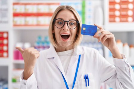 Photo for Young caucasian woman working at pharmacy drugstore holding credit card pointing thumb up to the side smiling happy with open mouth - Royalty Free Image