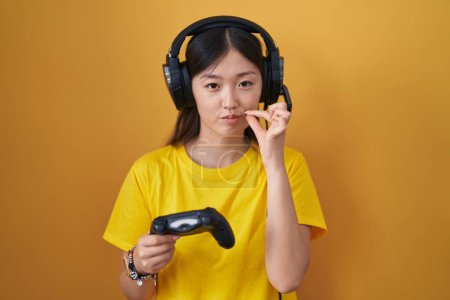 Téléchargez les photos : Jeune femme chinoise jouant jeu vidéo tenant la bouche du contrôleur et les lèvres fermées comme zip avec les doigts. secret et silencieux, tabou parlant - en image libre de droit