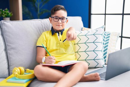 Téléchargez les photos : Jeune enfant hispanique faisant des devoirs assis sur le canapé pointant vers vous et la caméra avec les doigts, souriant positif et joyeux - en image libre de droit