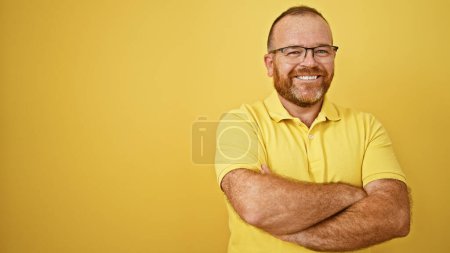 Alegre guapo hombre caucásico de mediana edad exudando confianza y alegría, de pie con una divertida expresión de risa, sus brazos cruzados sobre un fondo aislado amarillo vibrante