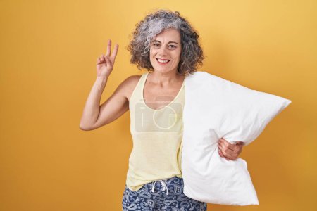 Téléchargez les photos : Femme d'âge moyen avec les cheveux gris portant un pyjama étreignant oreiller souriant en regardant la caméra montrant les doigts faisant signe de victoire. numéro deux. - en image libre de droit