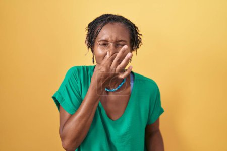 Téléchargez les photos : Femme africaine avec dreadlocks debout sur fond jaune sentant quelque chose de puant et dégoûtant, odeur intolérable, retenant souffle avec les doigts sur le nez. mauvaise odeur - en image libre de droit