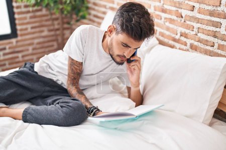 Photo for Young hispanic man talking on smartphone writing on book at bedroom - Royalty Free Image