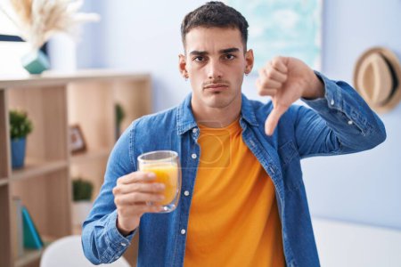 Téléchargez les photos : Bel homme hispanique boire un verre de jus d'orange avec le visage en colère, signe négatif montrant aversion avec les pouces vers le bas, concept de rejet - en image libre de droit
