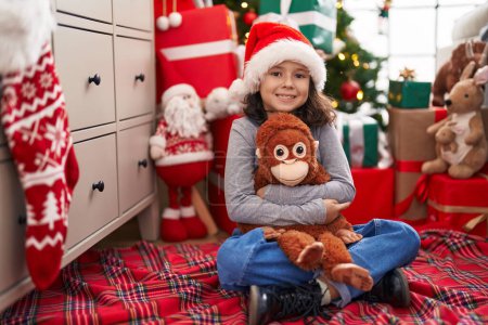 Photo for Adorable chinese girl hugging monkey doll sitting on floor by christmas tree at home - Royalty Free Image