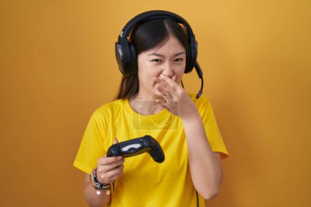Téléchargez les photos : Jeune femme chinoise jouant le contrôleur de tenue de jeu vidéo sentant quelque chose puant et dégoûtant, odeur intolérable, retenant souffle avec les doigts sur le nez. mauvaise odeur - en image libre de droit