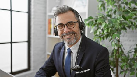 Téléchargez les photos : Homme d'affaires hispanique confiant, jeune et aux cheveux gris, souriant et travaillant au bureau avec son ordinateur portable, à l'écoute du son du succès grâce à son casque. - en image libre de droit