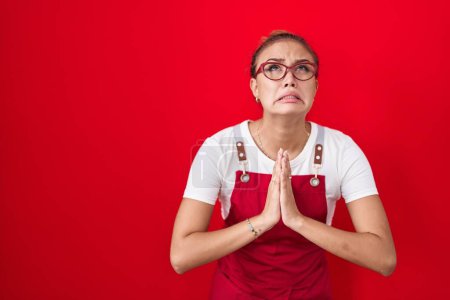 Photo for Young hispanic woman wearing waitress apron over red background begging and praying with hands together with hope expression on face very emotional and worried. begging. - Royalty Free Image