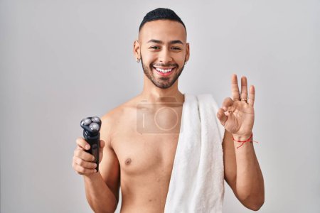 Foto de Joven hombre hispano sosteniendo navaja eléctrica haciendo signo de ok con los dedos, sonriendo gesto amistoso excelente símbolo - Imagen libre de derechos
