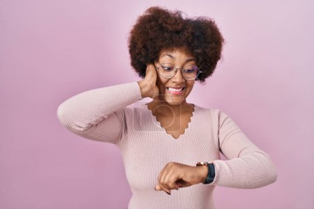 Téléchargez les photos : Jeune femme afro-américaine debout sur fond rose regardant le temps de la montre inquiet, peur de se faire tard - en image libre de droit