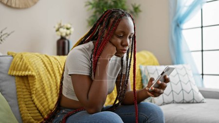 Photo for Beautiful african american woman, relaxed at home, intensely concentrates on typing a message on her phone as she sits on the sofa. - Royalty Free Image