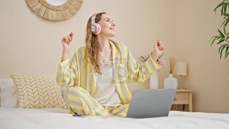 Téléchargez les photos : Jeune femme blonde écoutant de la musique assise sur le lit dansant dans la chambre - en image libre de droit
